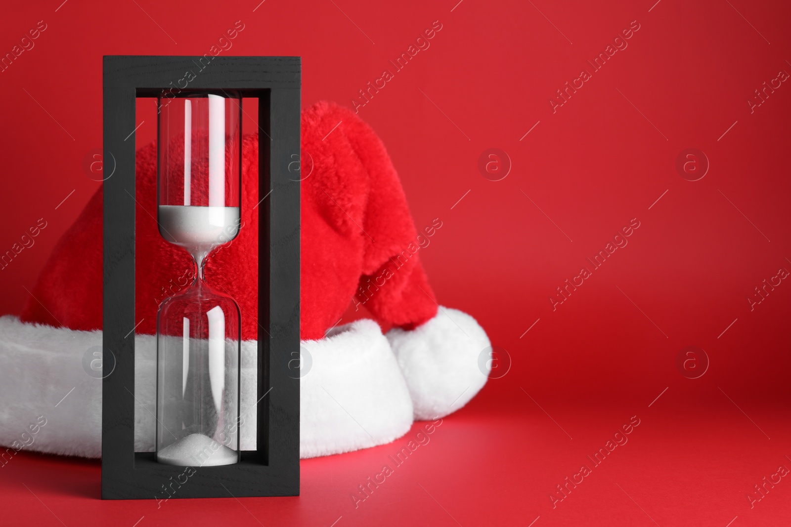 Photo of Hourglass and Santa hat on red background, space for text. Christmas countdown