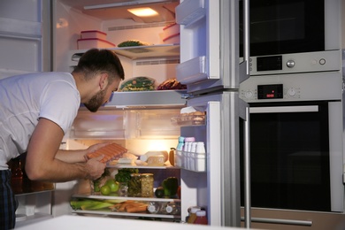Photo of Young man looking for food in refrigerator at night