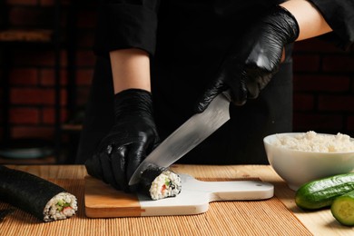 Photo of Chef in gloves cutting sushi roll at wooden table, closeup
