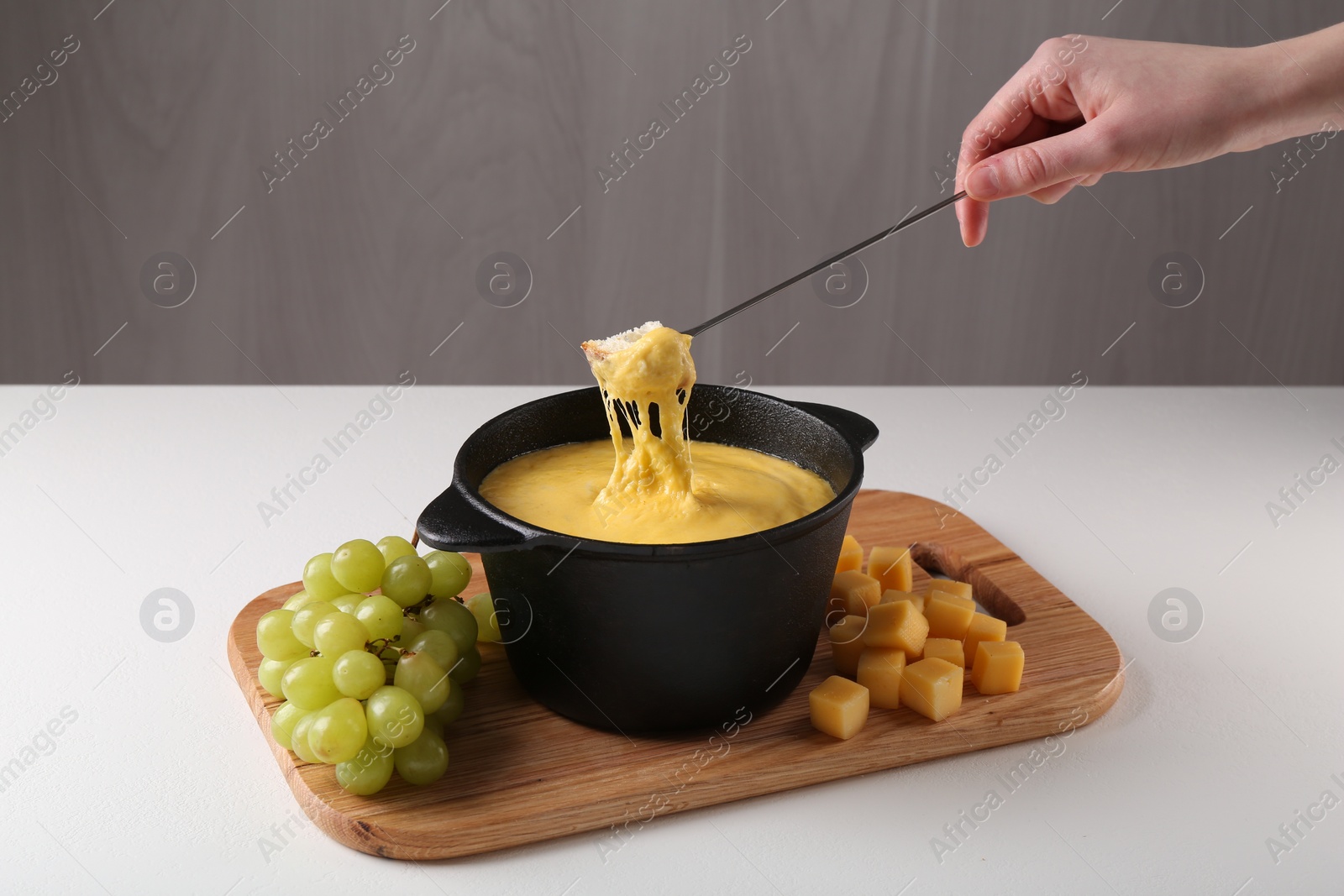 Photo of Woman dipping piece of bread into fondue pot with tasty melted cheese at white table, closeup