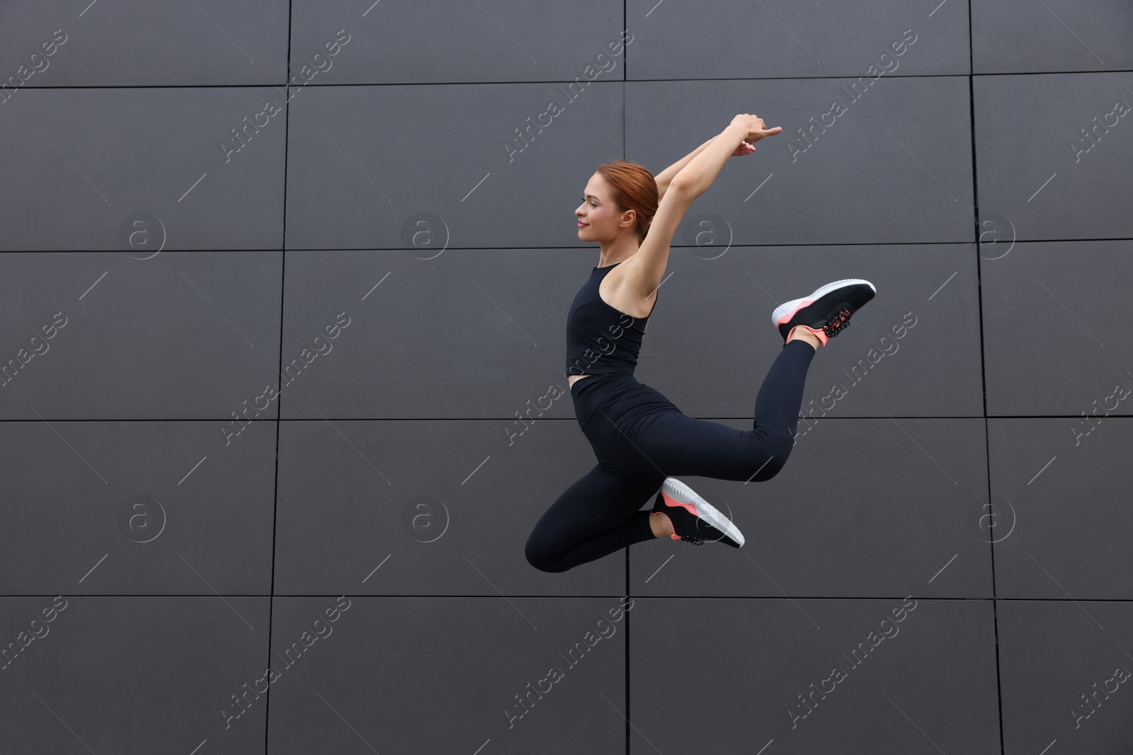 Photo of Beautiful woman in gym clothes jumping near dark grey wall on street, space for text