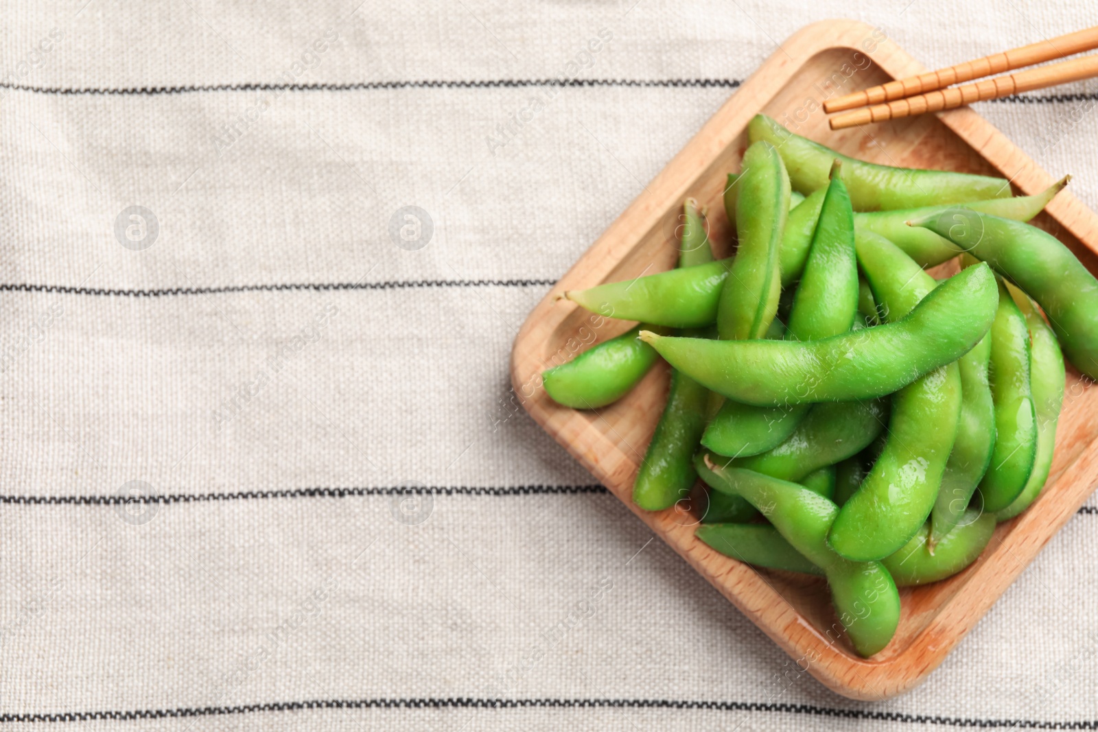 Photo of Wooden plate with green edamame beans in pods and chopsticks on towel, top view. Space for text