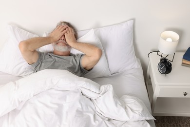 Photo of Senior man covering face in bed and alarm clock on bedside table at home
