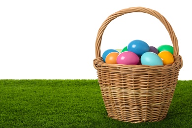 Photo of Wicker basket with bright painted Easter eggs on green grass against white background. Space for text