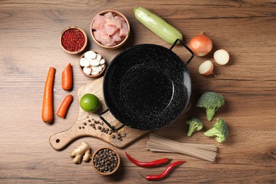 Photo of Empty iron wok surrounded by raw ingredients on wooden table, flat lay