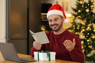 Celebrating Christmas online with exchanged by mail presents. Man reading greeting card during video call on laptop at home