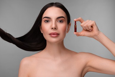 Beautiful young woman with long healthy hair holding ampoule on grey background