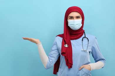 Photo of Muslim woman in hijab, medical uniform and protective mask pointing at something on light blue background. Space for text