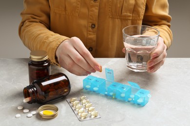 Woman putting pill into weekly organizer at grey marble table, closeup