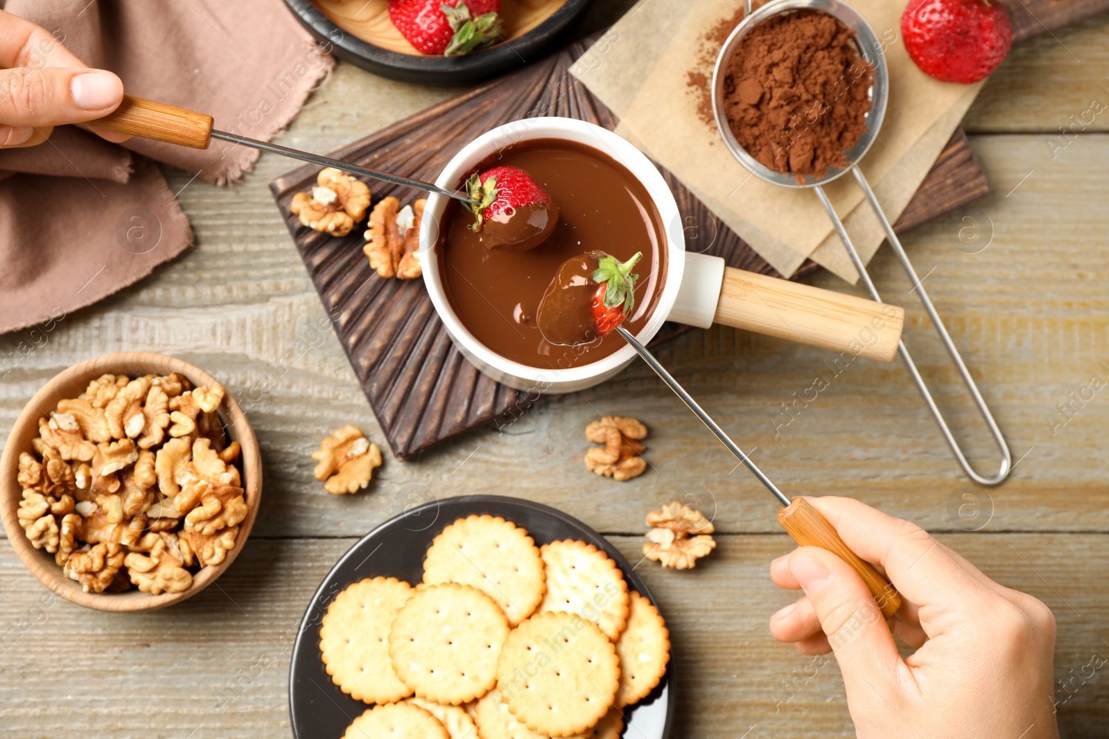 Photo of People dipping strawberry into fondue pot with milk chocolate on wooden table, top view