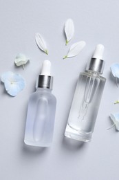 Bottles of cosmetic serums and beautiful flowers on light grey background, flat lay