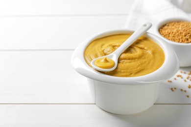 Photo of Spoon and bowl of tasty mustard sauce on white wooden table, space for text