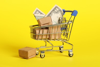 Photo of Small metal shopping cart with boxes and dollar bill on yellow background