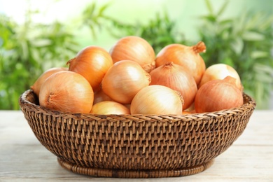 Photo of Wicker basket with ripe onions on white wooden table against blurred background