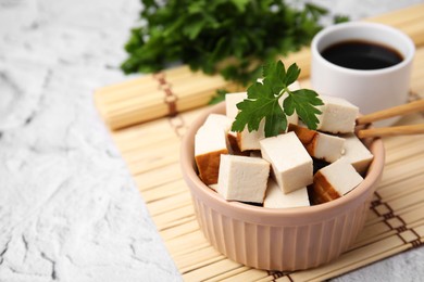 Photo of Bowl of smoked tofu cubes, soy sauce and parsley on white textured table, closeup. Space for text