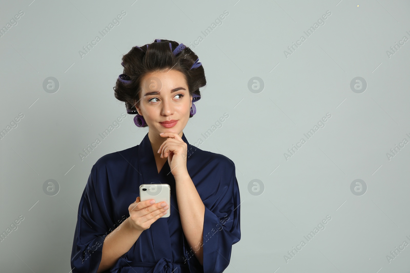 Photo of Happy young woman in silk bathrobe with hair curlers using smartphone on grey background, space for text