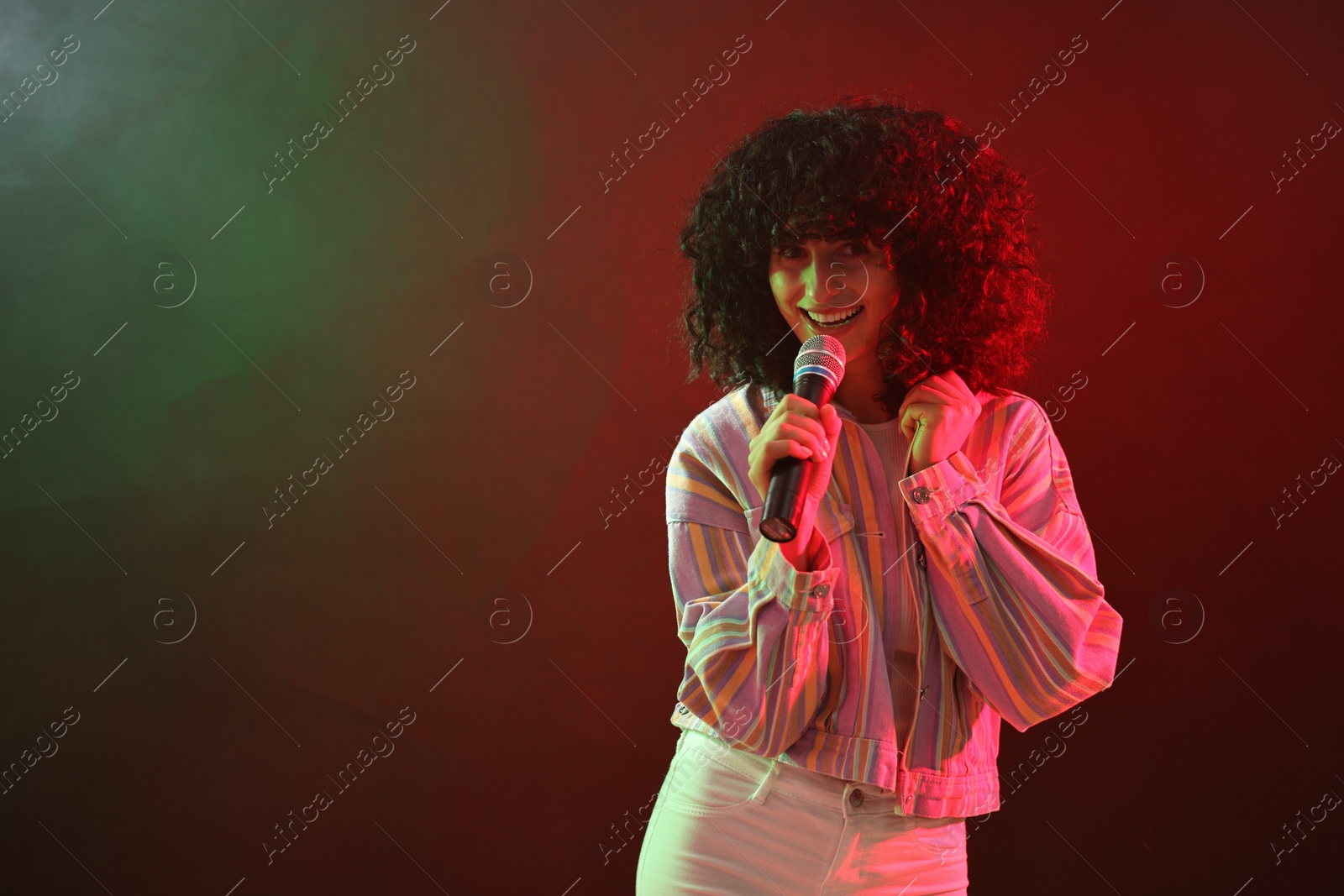 Photo of Beautiful young woman with microphone singing on color background in neon lights. Space for text