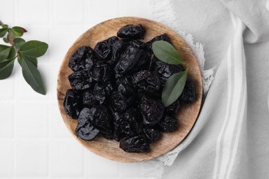 Photo of Sweet dried prunes and green leaves on white tiled table, top view