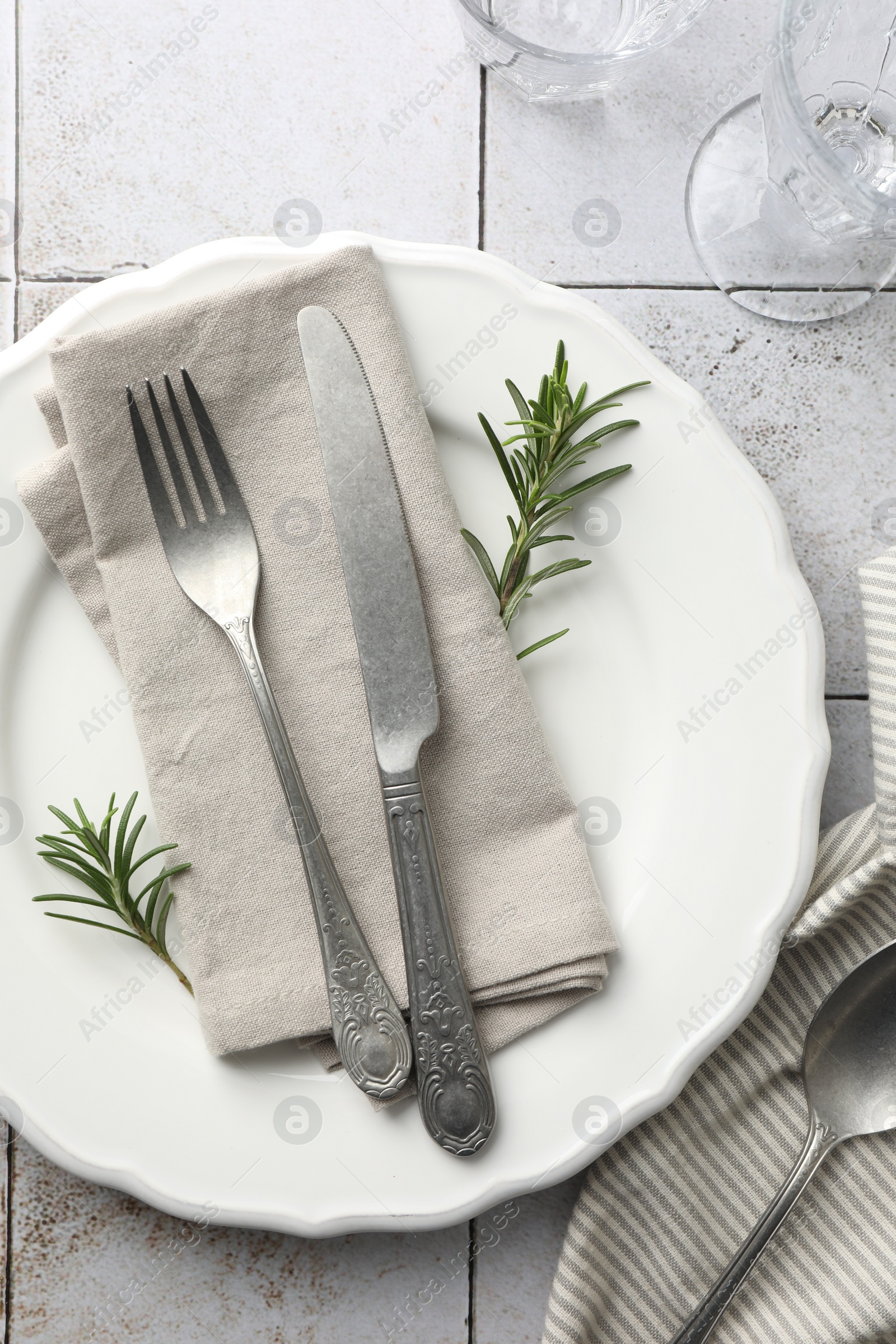 Photo of Stylish setting with cutlery, napkin, rosemary and plate on light tiled table, top view