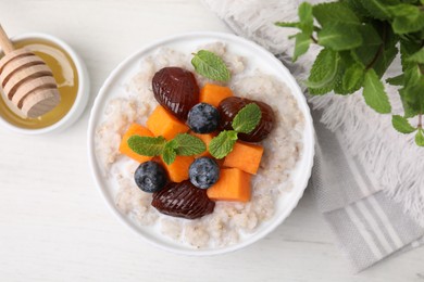 Photo of Delicious barley porridge with blueberries, pumpkin, dates and mint served with honey on white table, flat lay