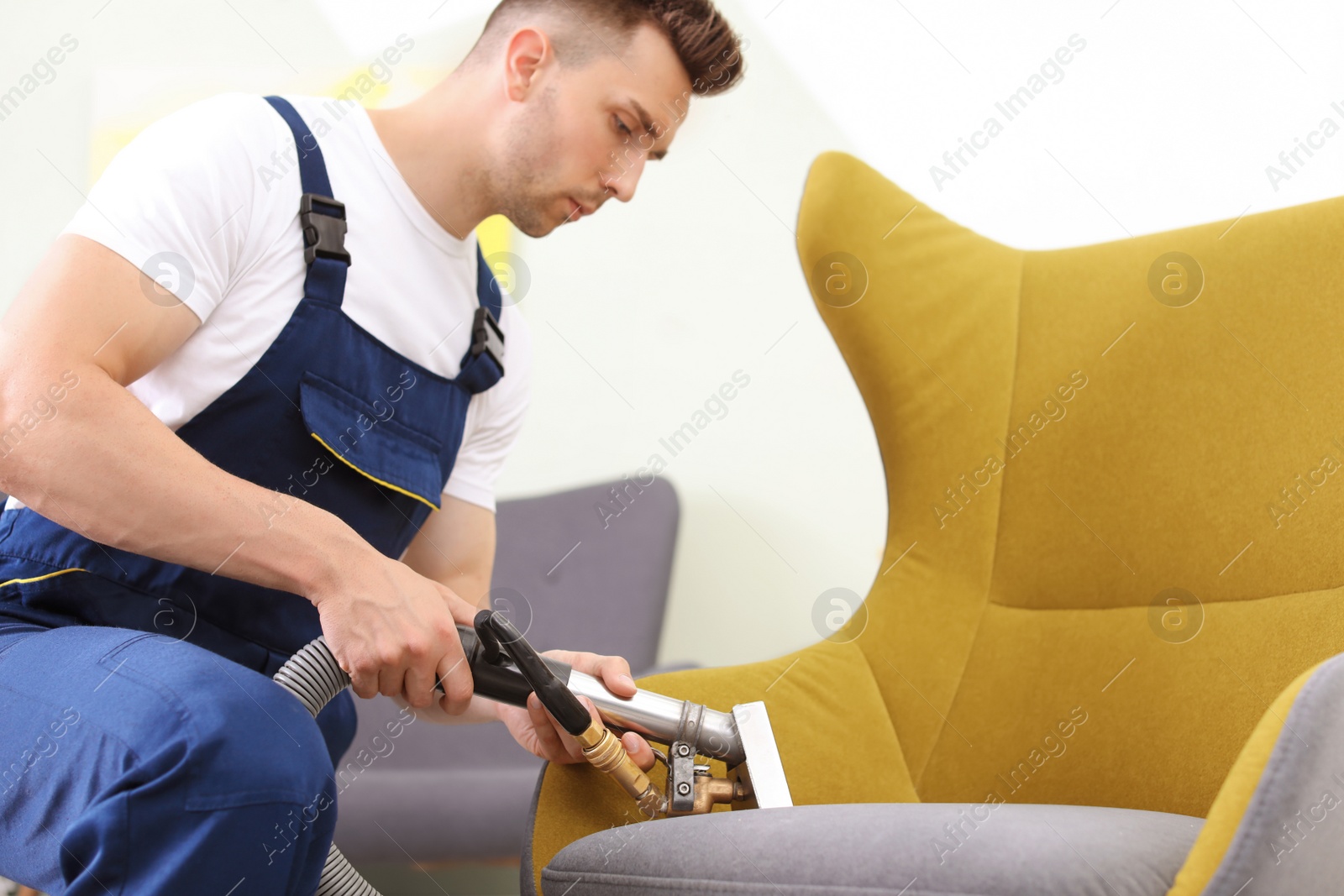 Photo of Male worker removing dirt from armchair with professional vacuum cleaner indoors