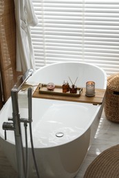 Wooden tray with cosmetic products, burning candles, and reed air freshener on bath tub in bathroom