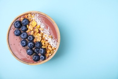 Photo of Bowl of delicious fruit smoothie with fresh blueberries, granola and coconut flakes on light blue background, top view. Space for text