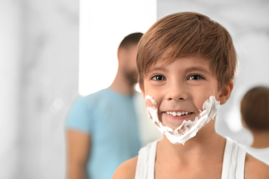 Photo of Happy little boy with shaving foam on face in bathroom, space for text. Dad and son having fun
