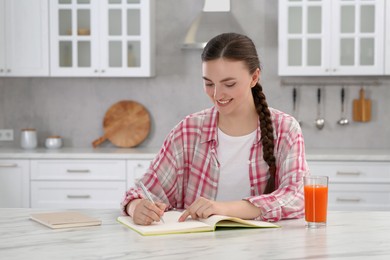 Beautiful young woman writing in notebook at white marble table indoors