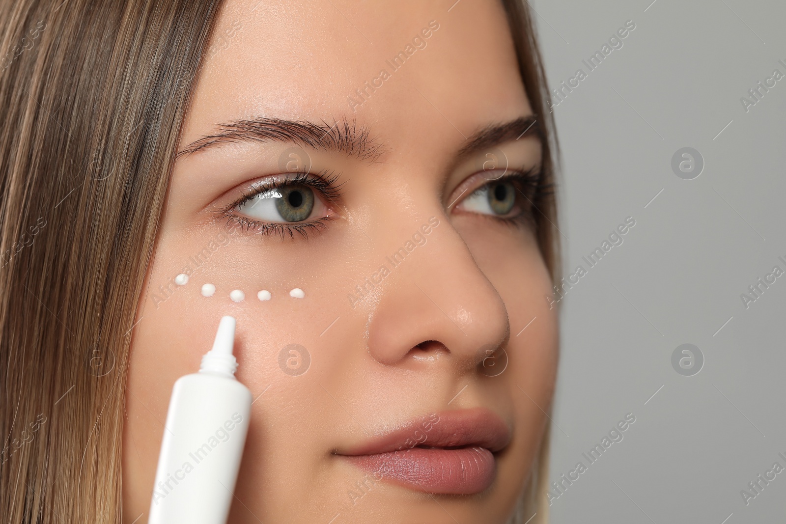 Photo of Young woman applying cream under eyes on light grey background, closeup