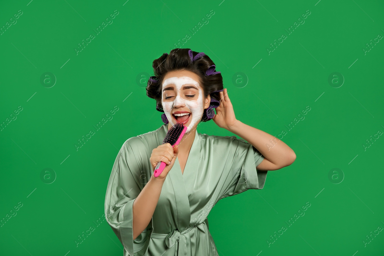 Photo of Happy young woman in silk bathrobe with hair curlers and facial mask singing into hairbrush on green background