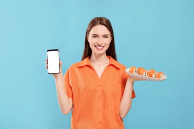 Happy young woman with plate of sushi rolls and smartphone on light blue background. Space for text