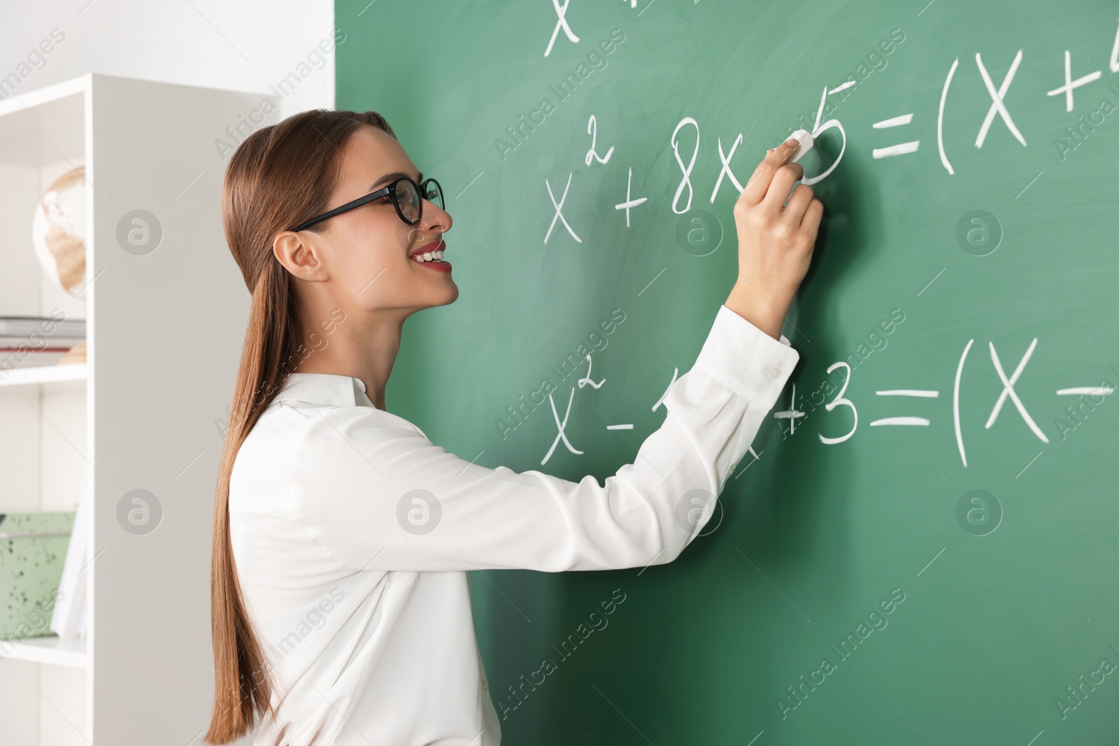 Photo of Young math’s teacher writing mathematical equations near chalkboard in classroom