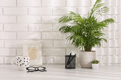 Photo of Stylish office workplace. Frame, plants, glasses, pens and cup on white table near brick wall