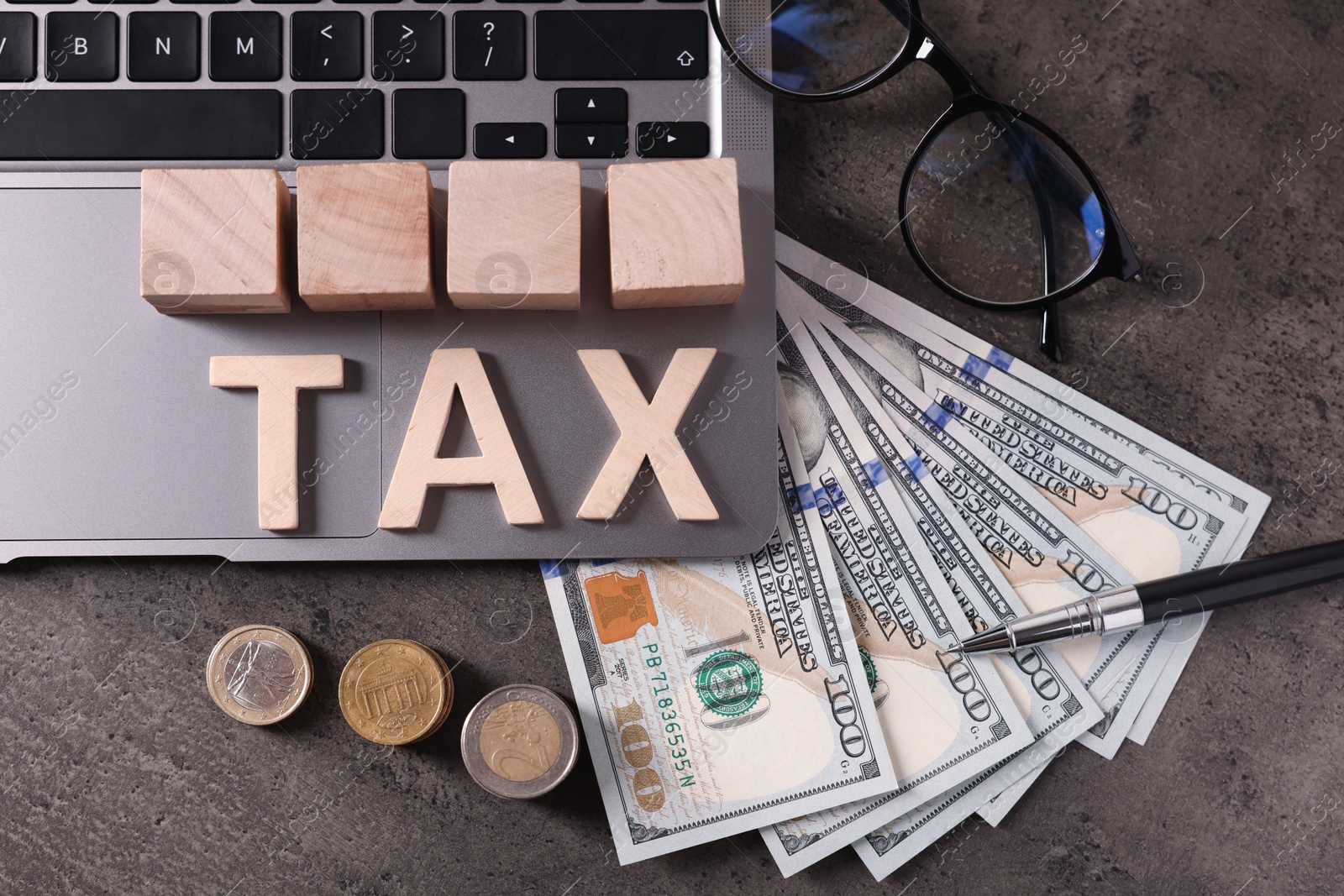 Photo of Word Tax made of wooden letters, cubes, laptop, banknotes and coins on grey table, flat lay