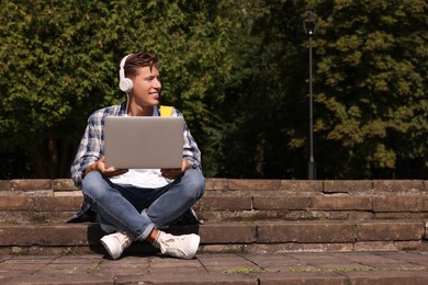 Photo of Happy young student with headphones studying with laptop on steps in park, space for text
