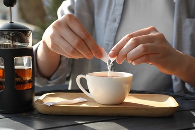 Woman adding sugar into cup of tea at black wooden table in outdoor cafe, closeup