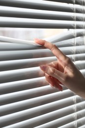 Photo of Woman separating slats of white blinds indoors, closeup