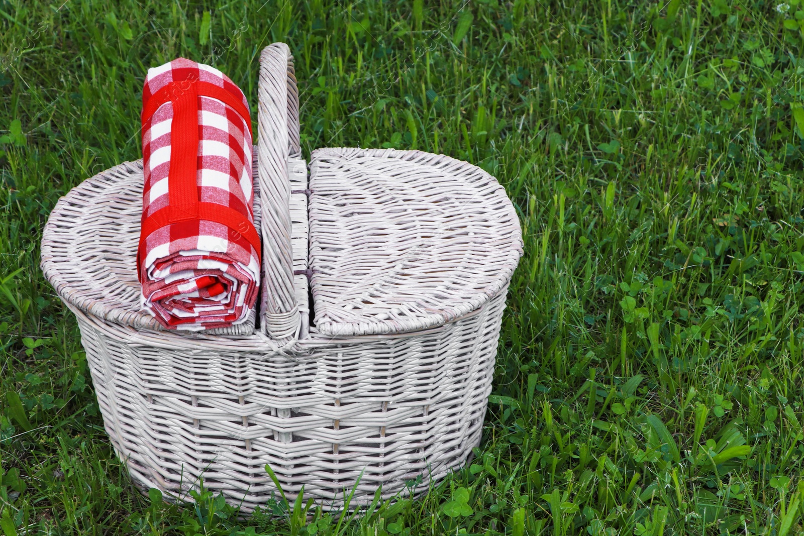 Photo of Rolled checkered tablecloth with picnic basket on green grass outdoors, space for text