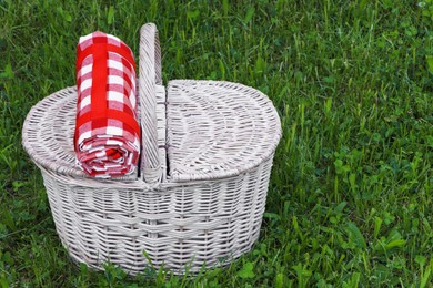 Rolled checkered tablecloth with picnic basket on green grass outdoors, space for text