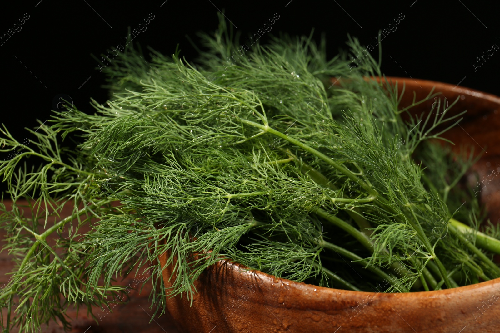 Photo of Wooden bowl of fresh green dill with water drops on table against black background, closeup