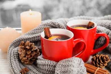 Cups of hot winter drink with knitted sweater on window sill indoors