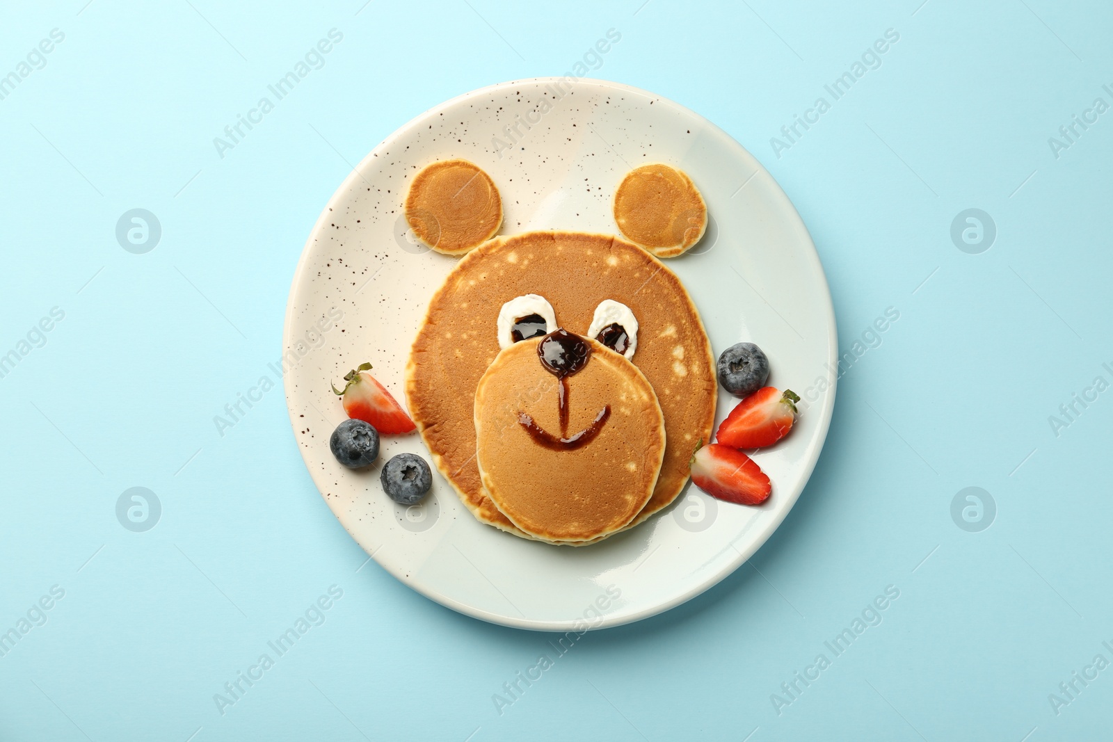 Photo of Creative serving for kids. Plate with cute bear made of pancakes and berries on light blue table, top view