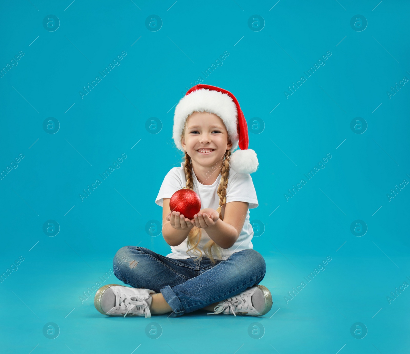 Photo of Cute little child wearing Santa hat holding Christmas decoration on blue background