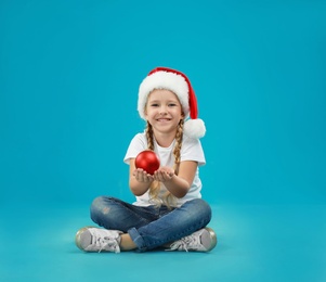 Photo of Cute little child wearing Santa hat holding Christmas decoration on blue background