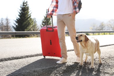 Photo of Man with red suitcase and adorable dog near road, closeup. Traveling with pet