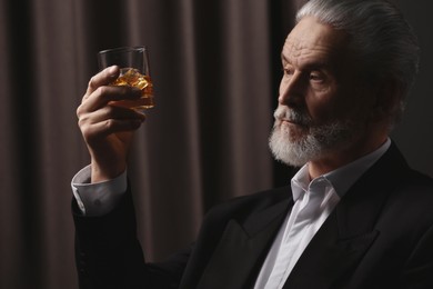 Photo of Senior man in formal suit holding glass of whiskey with ice cubes on brown background