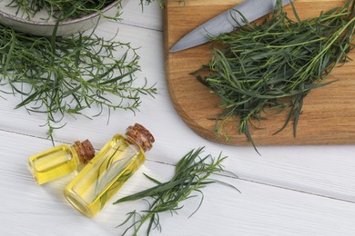 Photo of Bottles of essential oil and fresh tarragon sprigs on white wooden table, above view