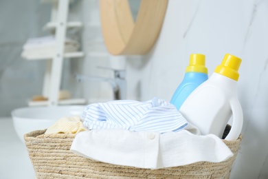 Photo of Bottles of detergent and children's clothes in wicker basket indoors, closeup