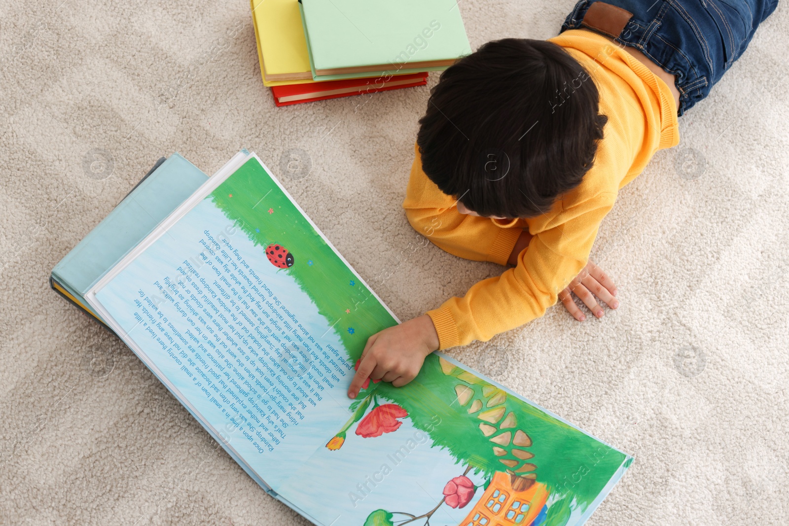 Photo of Cute little boy reading book on floor at home, above view